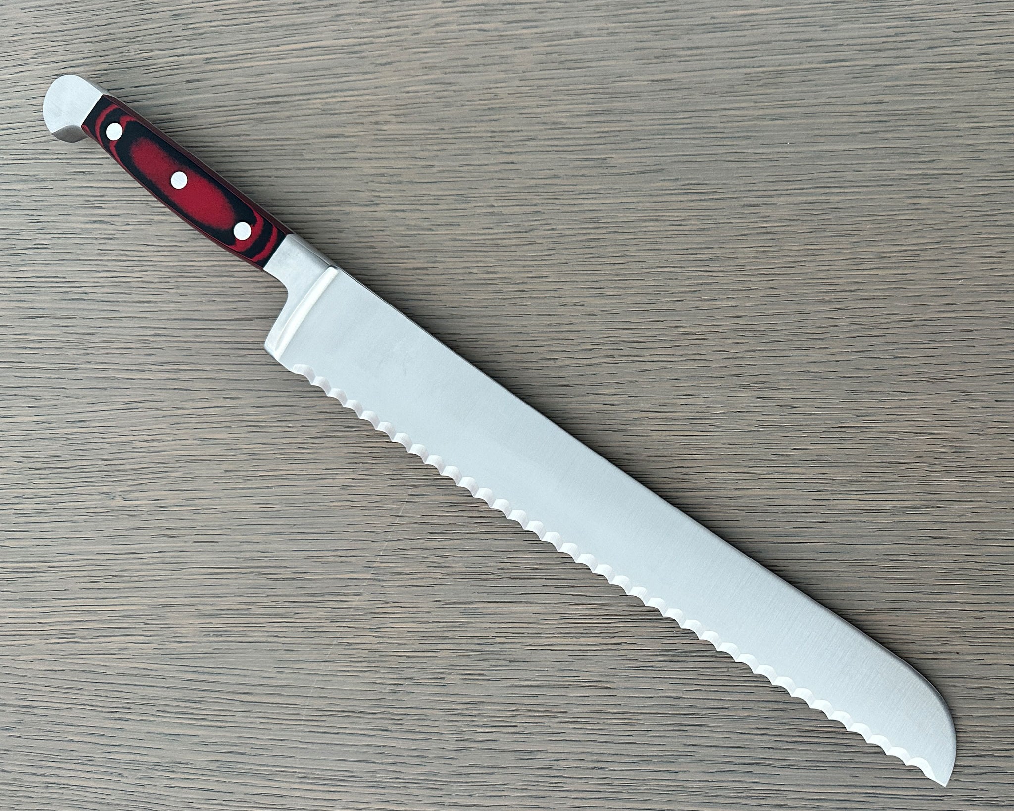 A Güde Solingen serrated bread knife reverse side of wavy-edge blade and an ergonomic red-and-black Micarta handle, resting diagonally on a gray wood-grain board