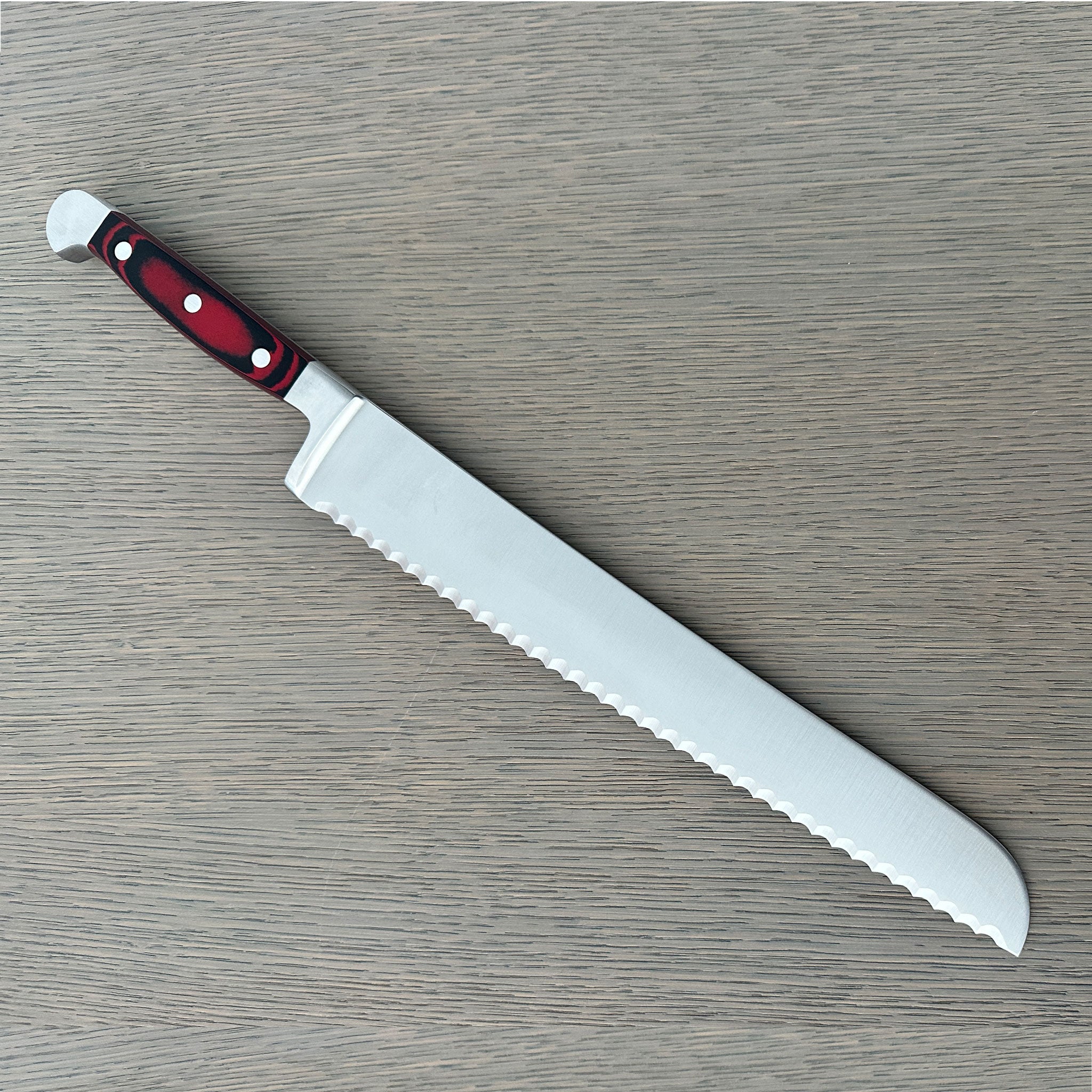 A Güde Solingen serrated bread knife reverse side of wavy-edge blade and an ergonomic red-and-black Micarta handle, resting diagonally on a gray wood-grain board