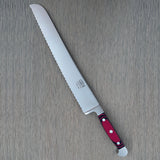 A Güde Solingen serrated bread knife with a wavy-edge blade and an ergonomic red-and-black Micarta handle, resting diagonally on a gray wood-grain board