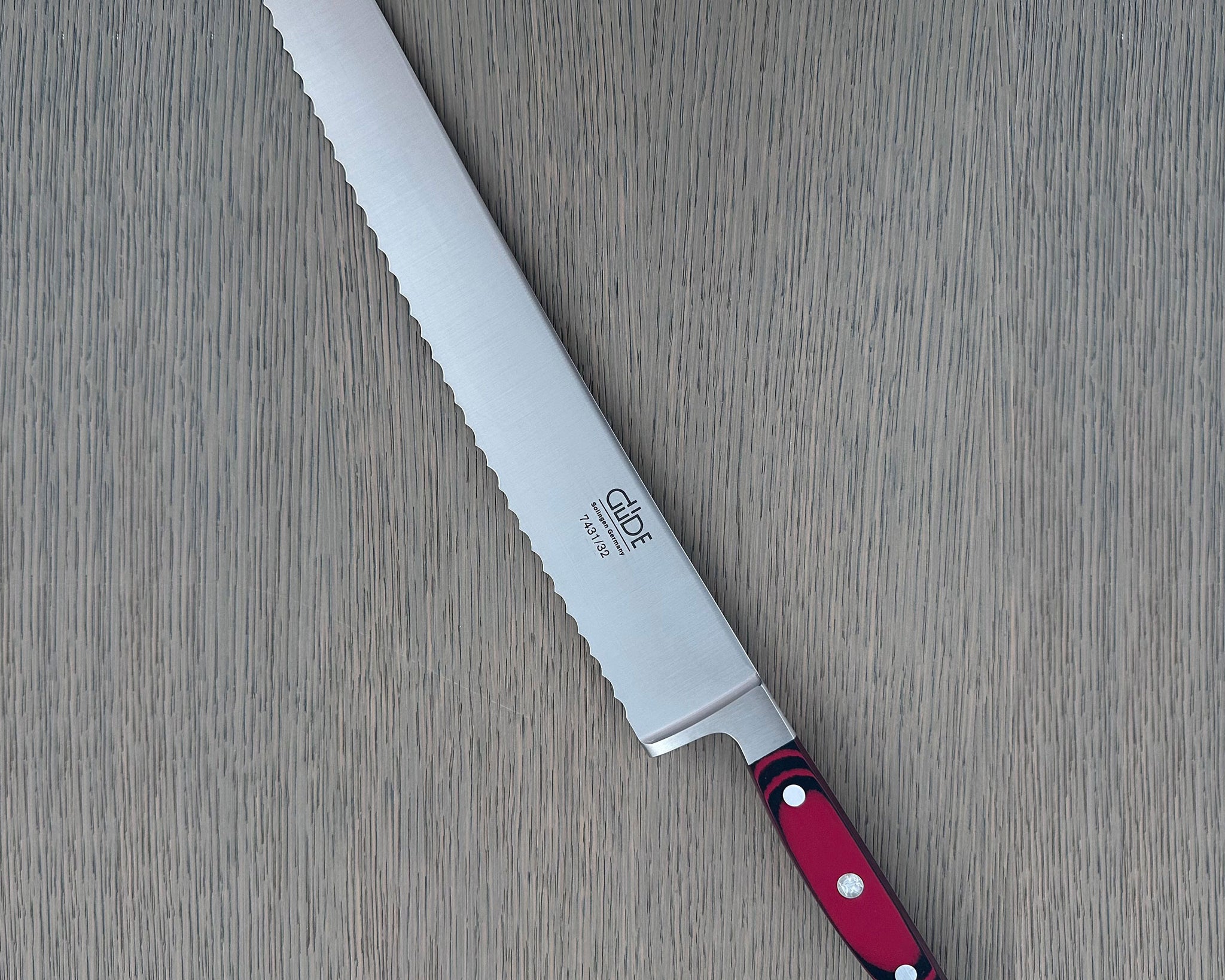A Güde Solingen serrated bread knife with a wavy-edge blade and an ergonomic red-and-black Micarta handle, resting diagonally on a gray wood-grain board