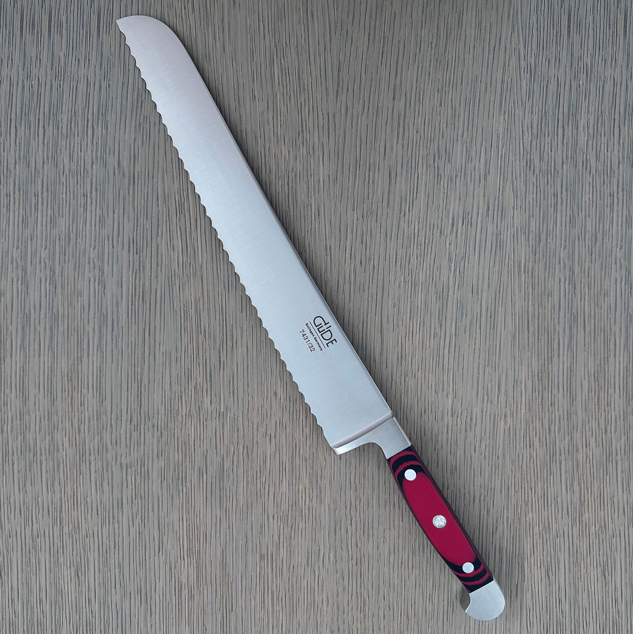 A Güde Solingen serrated bread knife with a wavy-edge blade and an ergonomic red-and-black Micarta handle, resting diagonally on a gray wood-grain board