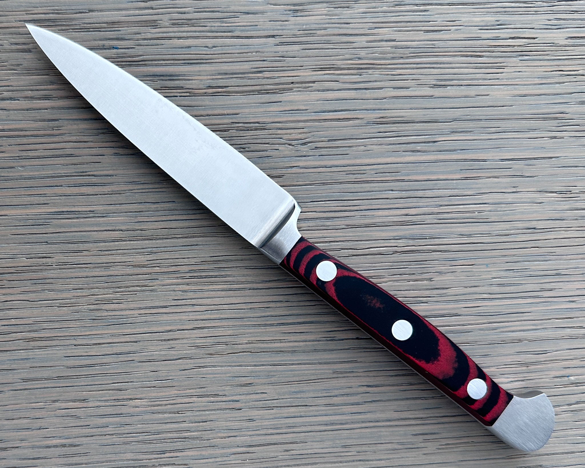 A Gude Micarta paring knife, revserse side with steel blade and a marbled red, black handle displayed diagonally on a light gray wood-grain surface.