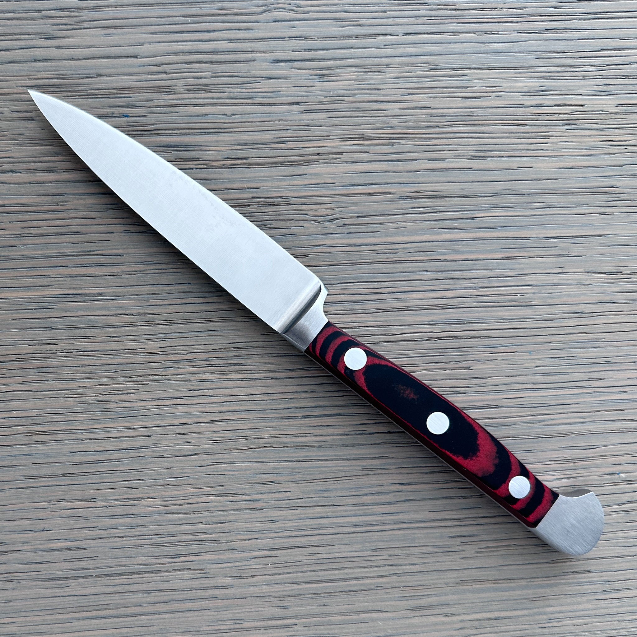 A Gude Micarta paring knife, revserse side with steel blade and a marbled red, black handle displayed diagonally on a light gray wood-grain surface.