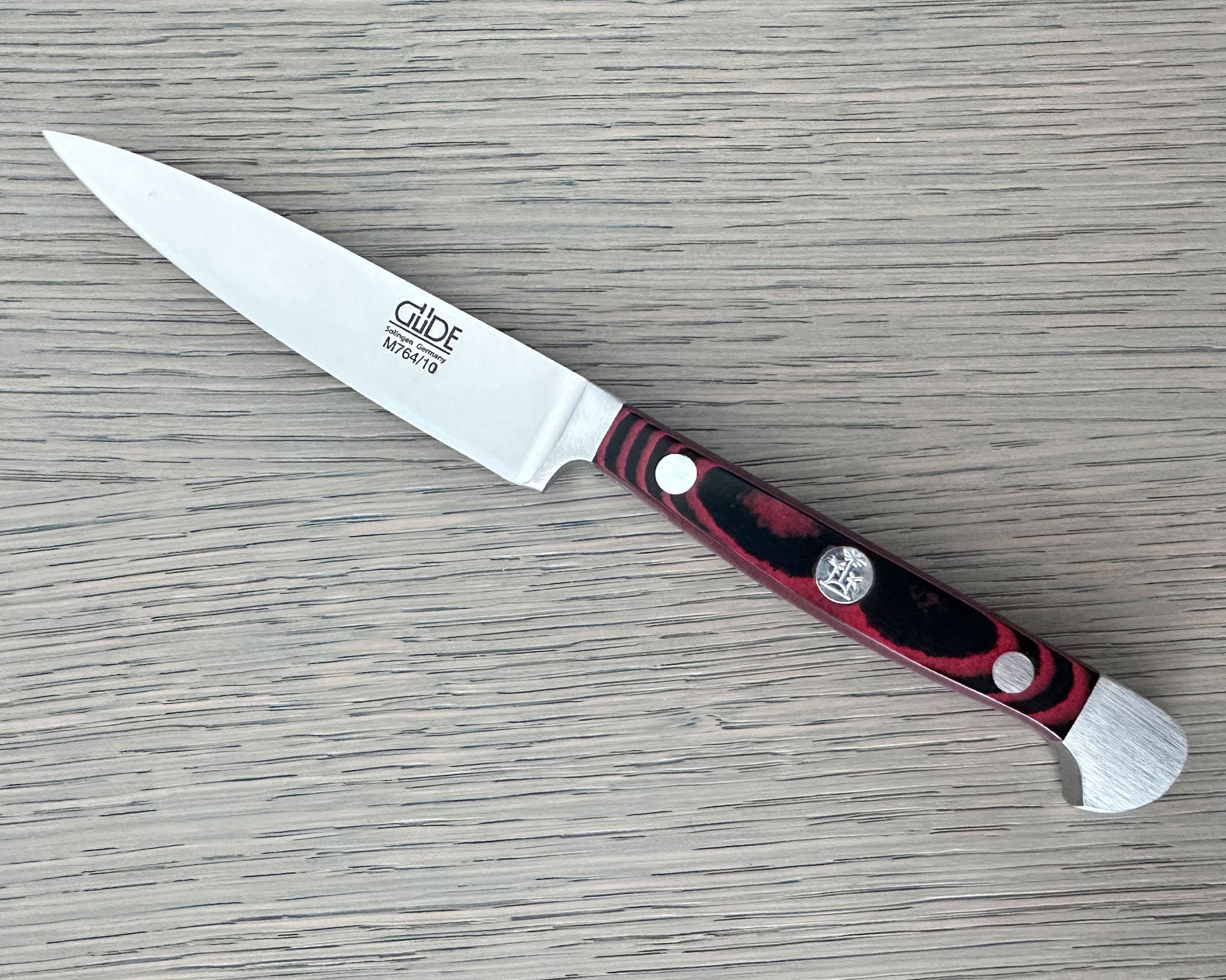 A Gude Micarta paring knife with steel blade and a marbled red, black handle displayed diagonally on a light gray wood-grain surface.
