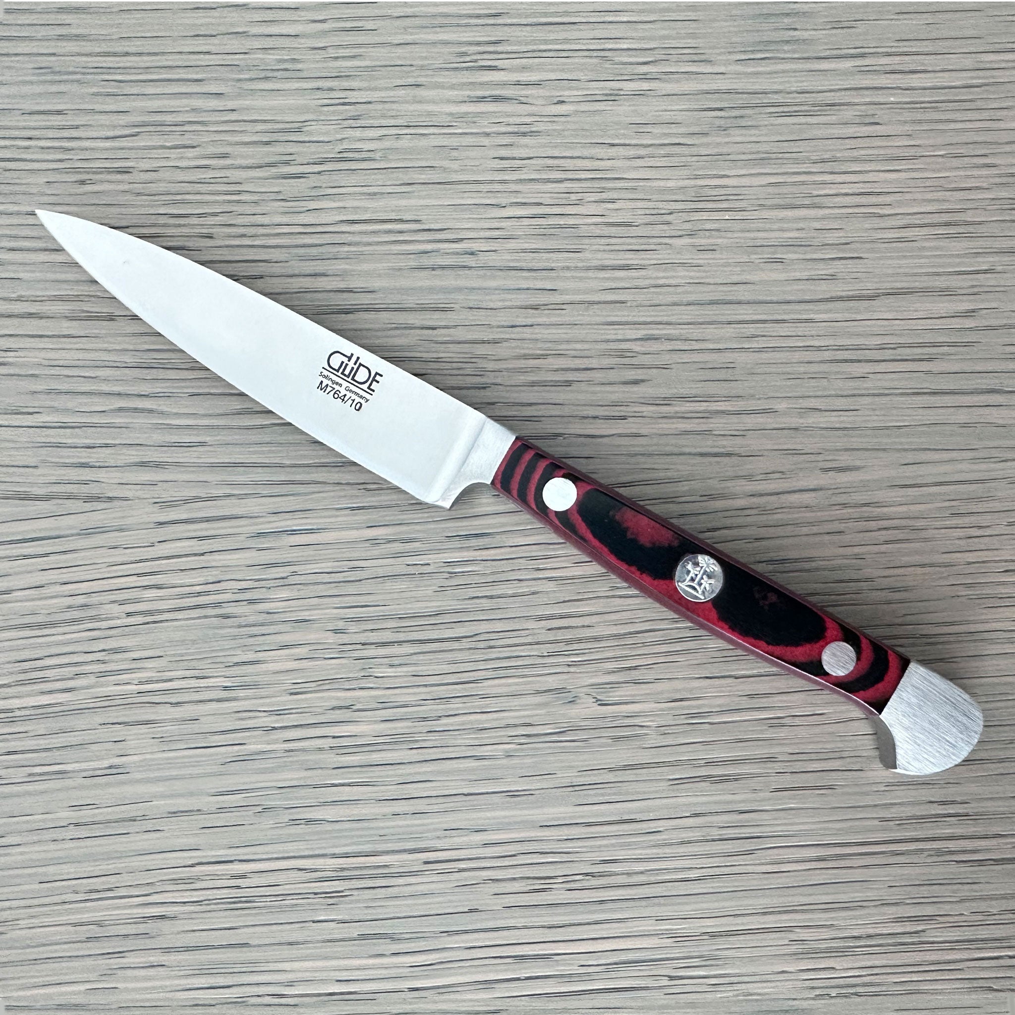 A Gude Micarta paring knife with steel blade and a marbled red, black handle displayed diagonally on a light gray wood-grain surface.
