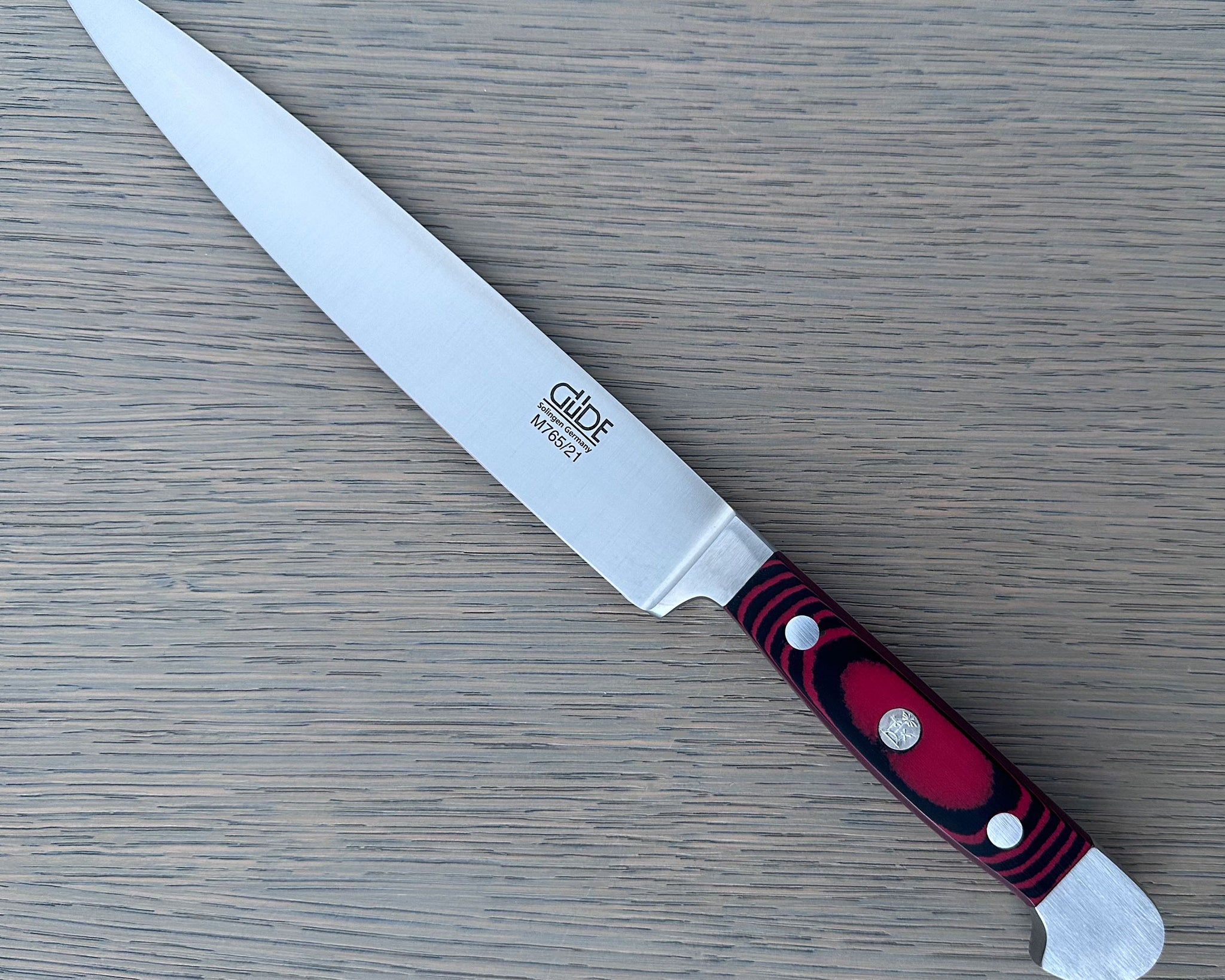 A Gude Micarta Slicing knife with steel blade and a marbled red, black handle displayed diagonally on a light gray wood-grain surface.