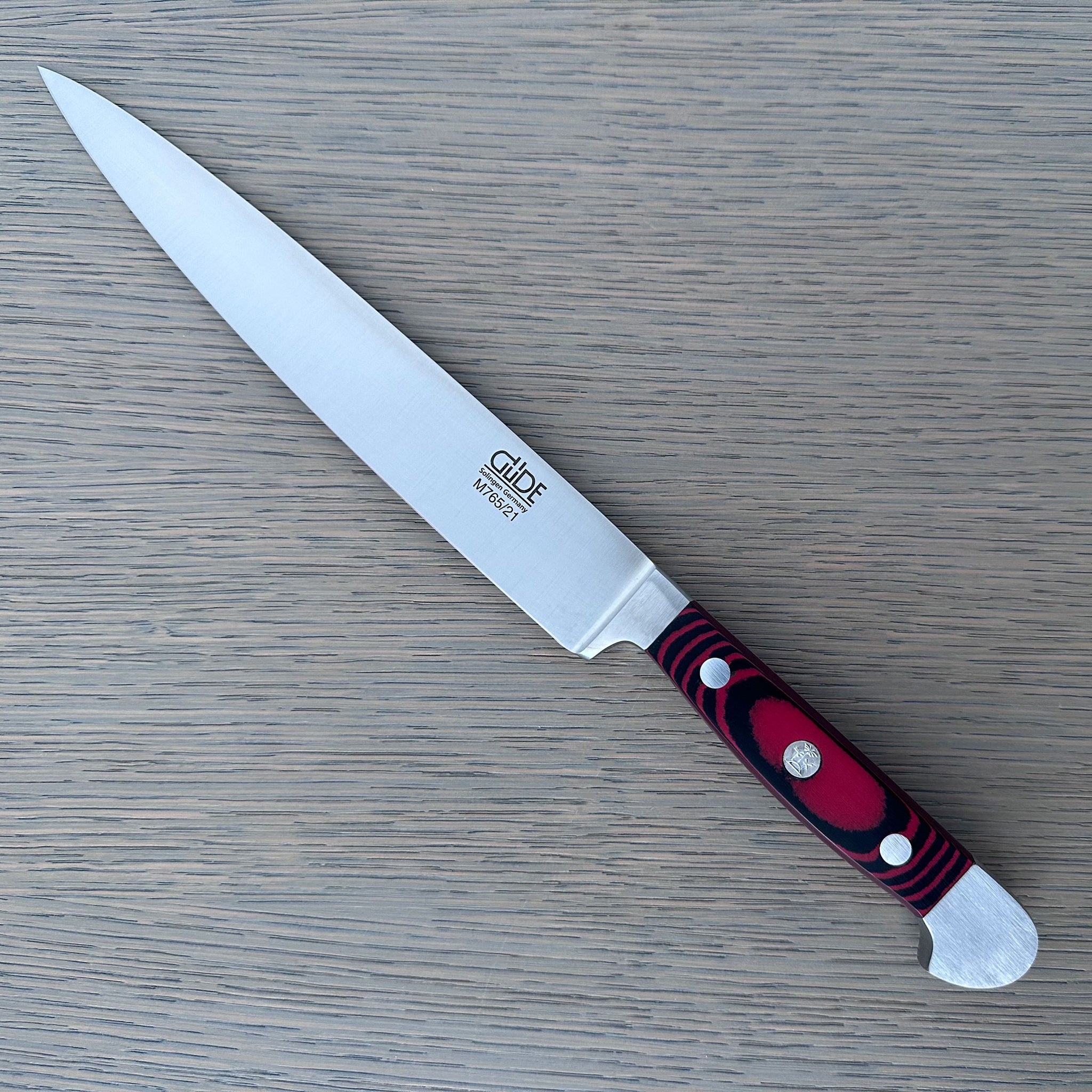 A Gude Micarta Slicing knife with steel blade and a marbled red, black handle displayed diagonally on a light gray wood-grain surface.